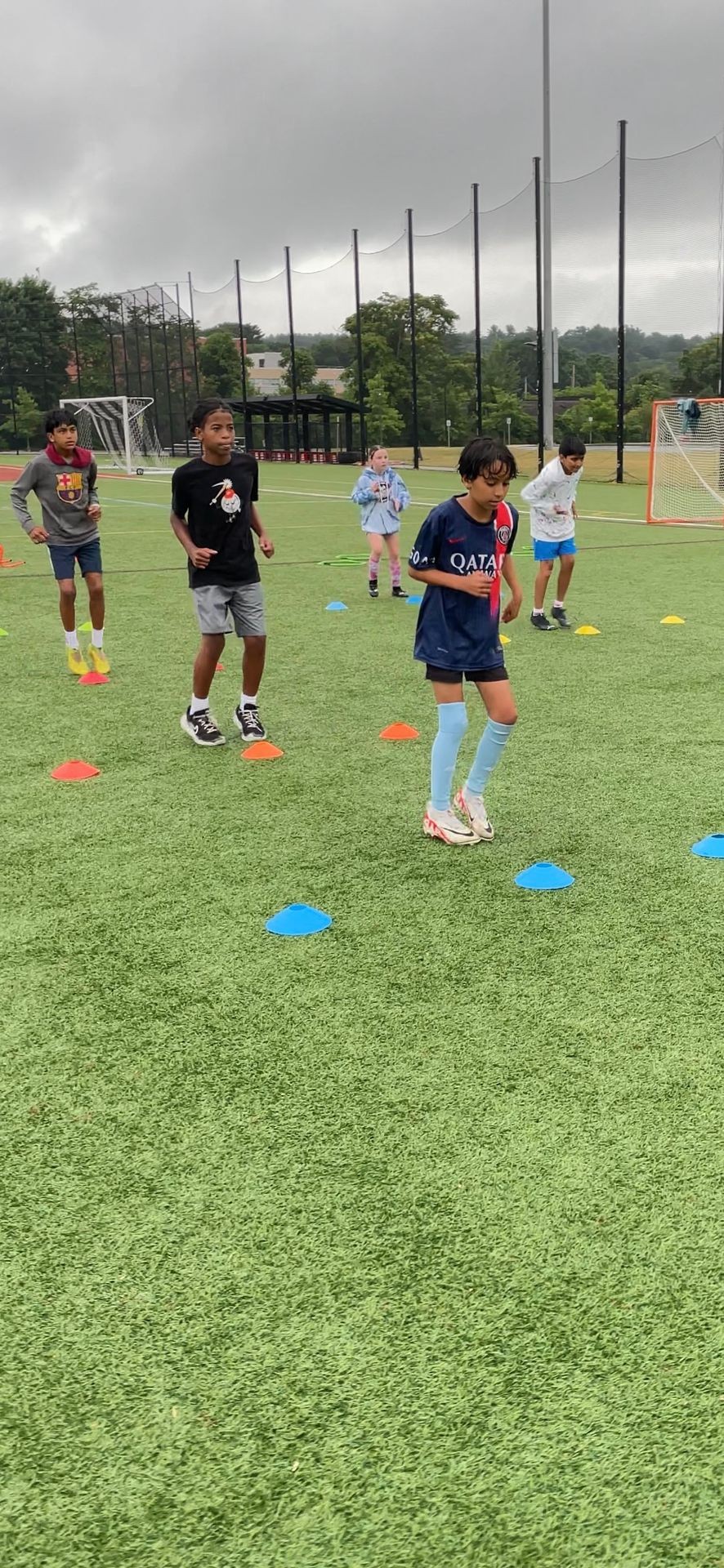 Children practicing drills on a soccer field, running between colorful cones under a cloudy sky.